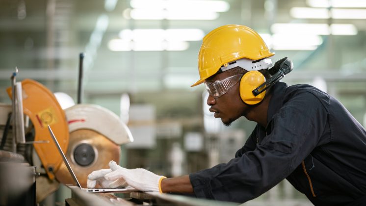Maintenance technician in PPE using a laptop beside industrial equipment, capturing work order feedback during maintenance.