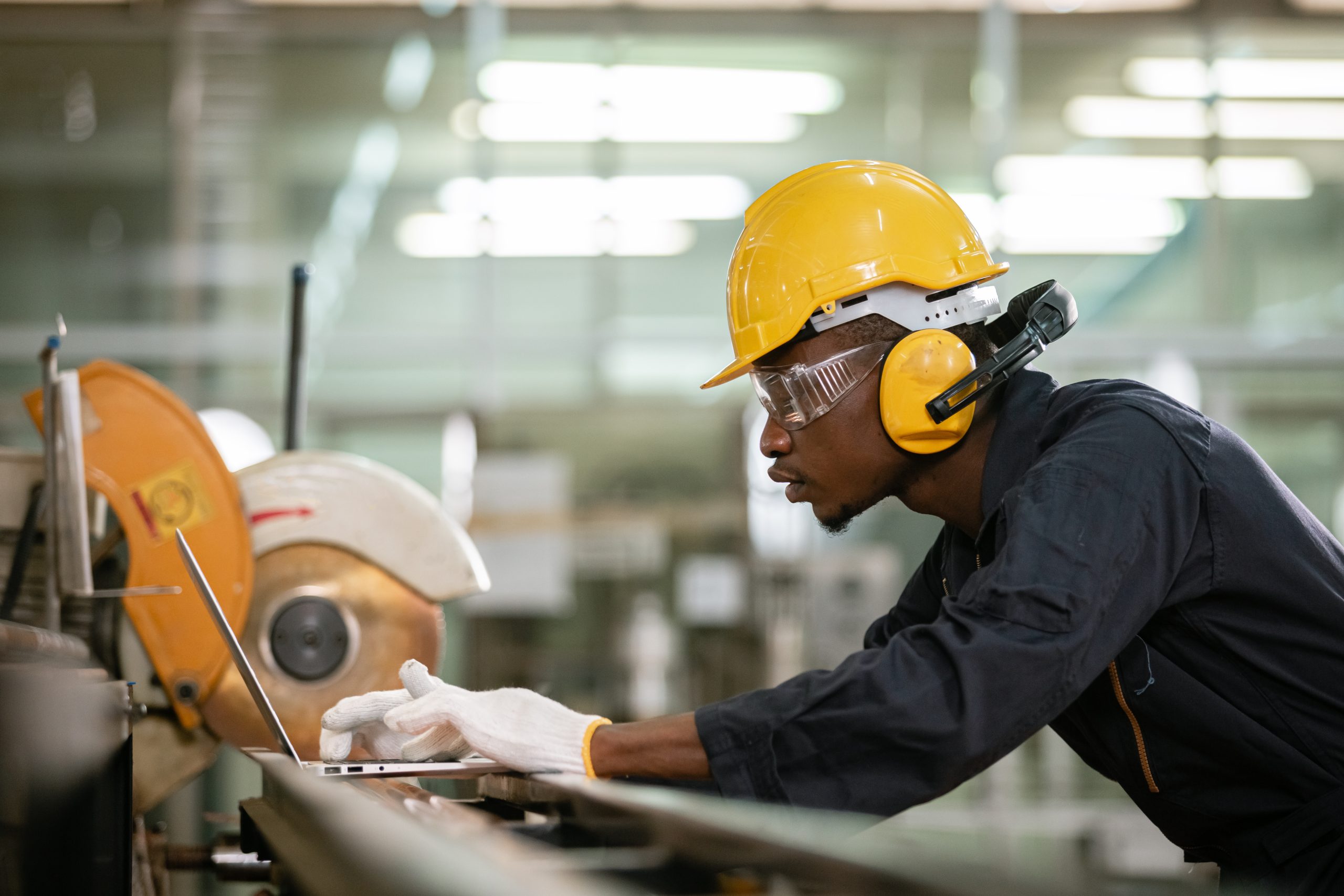 Maintenance technician in PPE using a laptop beside industrial equipment, capturing work order feedback during maintenance.